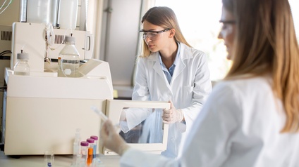 Zwei Personen in einem Laborkittel und mit einer Schutzbrille legen ein Fläschchen mit einer Probe für eine Analyse in ein Gerät, welches an einem Tisch im Labor steht.