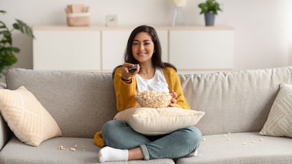 Eine Person sitzt im Schneidersitz auf einem hellen Sofa. Auf ihrem Schoß ist ein gelbliches Kissen, auf dem eine Schüssel, befüllt mit Popcorn ist. Die Person hält in der rechten Hand eine Fernbedienung und blickt geradeaus
