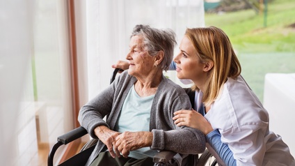 Neben einem großen Fenster sitzt eine Person in einem Rollstuhl. Rechts neben der Person im Rollstuhl ist eine weitere Person auf selber Höhe. Beide blicken aus dem Fenster