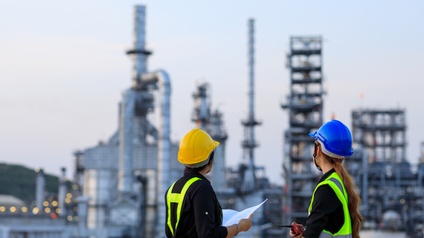 Rückenansicht zweier nebeneinander stehender Person mit Helm und Sicherheitswesten. Die rechte Person hält ein Walkie-Talkie. Im Hintergrund sind verschiedene Türme, Rohre und Leitungen aus metallenen Materialien. Die Personen blicken zu den Türmen