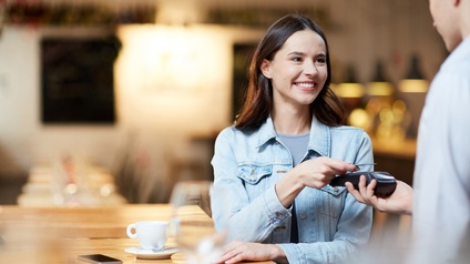 Frontalaufnahme einer seitlich an einem Tisch sitzenden Person, die in der rechten Hand ein Objekt hält, das sie über ein Gerät hält, das von einer anderen Person gehalten wird, deren Rücken im Bild ist. Vor der Person steht eine Tasse
