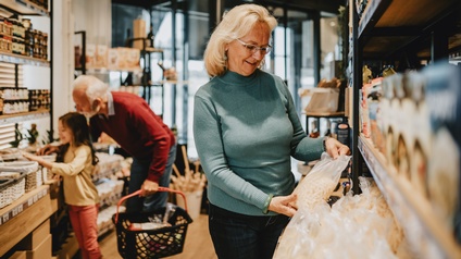 In einem Gang in einem Supermarkt steht rechts im Bild eine Person mit grauen Haaren und Brille, die ein Lebensmittelprodukt hält. Links stehen eine Person mit grauen Haaren und Bart sowie ein Kind. Die Person hält einen Einkaufskorb
