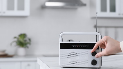 Ein Radio steht auf einem Tresen in einer hellen Küche. Die rechte Hand einer Person dreht an einem Rädchen des Radios
