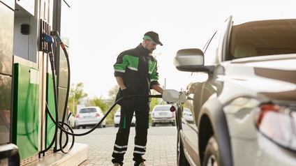 Eine Person in Arbeitskleidung steht an einem Auto neben einer Zapfsäule auf eine Tankstelle und tankt das Gefährt.