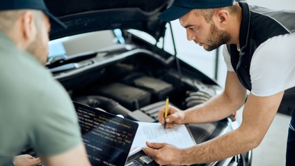 Zwei Personen mit Kappen beugen sich über geöffnete Motorhaube eines Fahrzeuges, eine Person nimmt auf Dokument Notizen vor, die andere Person betätigt aufgeklappten Laptop vor sich