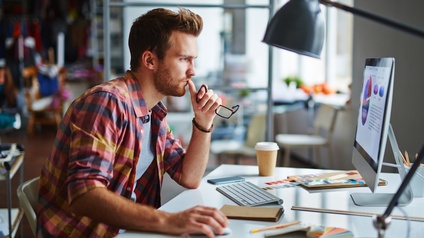 Seitliche Aufnahme einer an einem Tisch sitzenden Person, auf dem ein Bildschirm, eine Tastatur und andere Utensilien sind. Die Person hat ihre rechte Hand auf einer Computermaus und blickt auf den Bildschirm