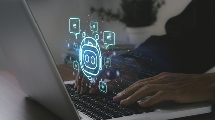 Eine Person tippt auf einem Laptop, der auf einem dunkelbraunen Holztisch steht. Vot dem Bildschirm sind Symbole zu sehen, ein Roboter in einem Kreis, rundherum Sprechblasen mit drei Strichen und leuchtende Punkte