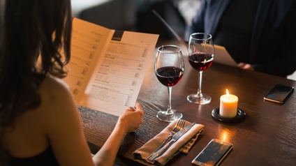 Zwei Weingläser gefüllt mit Rotwein auf Esstisch stehend, daneben entzundene Kerze, Smartphones und Serviette mit Besteck, am Tisch zwei Personen sitzend, die Speisekarten betrachten