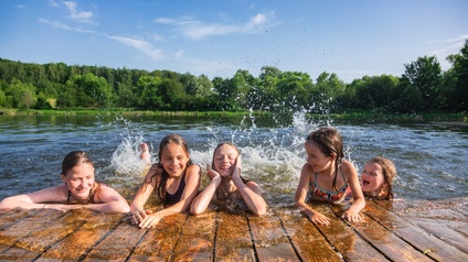 Lachende Kinder in einem See halten sich an einem Holzsteg  fest, im Hintergrund sind Bäume.