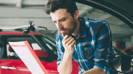 Portrait einer Person mit dunklen kurzen Haaren, Bart und kariertem Hemd, die konzentriert ein Dokument liest und sich mit einer Hand beim Bart abstützt, und mit der anderen Hand beim geöffneten Fenster eines Autos draußen anlehnt, im Hintergrund Autos