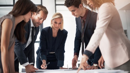 Fünf Personen in Bürokleidung stehen in einem Raum um einen Tisch. Auf dem Tisch liegen Zettel. Die Personen blicken auf die Zettel. Manche deuten darauf