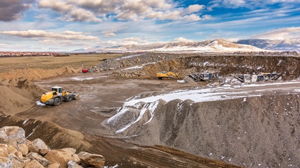 Gelber Bagger, der Gestein in einem teilweise verschneiten Tagebau bewegt, Hügel aus aufgeschüttetem Bergreich, im Hintergrund verschneite Berge und bewölkter Himmel