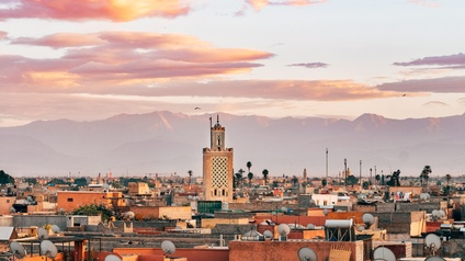 Panoramaaufnahme Marrakeschs mit den Dächern der Häuser sowie einem Minarett. Im Hintergrund ist die Gebirgsgruppe Atlas