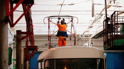 Ein Straßenbahnwagon befindet sich in einer Halle zur Wartung. Auf dem Dach des Wagons ist in Rückenansicht einer Person zu sehen. Die Person trägt Arbeitskleidung und arbeitet an einem Stromabnehmer.