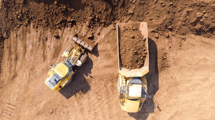 Aufnahme eines Baggers sowie eines Fahrzeuges mit einem großen Container aus der Vogelperspektive. Im Container ist Erde genauso wie auf der Schaufel des Baggers. Neben Bagger und Fahrzeug ist ein großer Haufen Erde