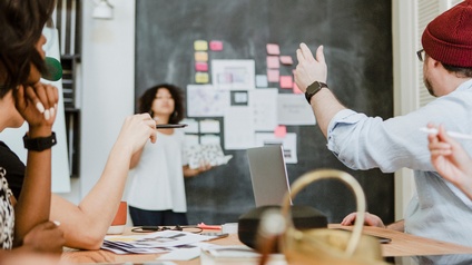 Um einen Tisch sitzen fünf Personen. Auf dem Tisch sind verschiedene Zettel, ein Tablet, ein Laptop und Tassen. Im Hintergrund steht eine Person neben einer Wand, auf der verschiedene Zettel kleben. Die am Tisch sitzenden Personen blicken zu dieser Person
