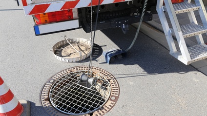 Neben einem Kanaldeckel steht ein Fahrzeug mit einer Trittbrettleiter hinten dran. Vom Fahrzeug hängen zwei Schnüre in den Kanaldeckel hinein