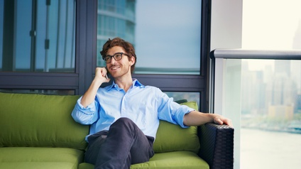 Eine Person in einem Hemd, mit Brille, dunklen Haaren und Bart sitzt auf einer Terrasse auf einem Sofa und telefoniert. Rechts neben der Person ist eine Glasreling