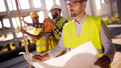 Es sind Personen abgebildet, eine davon im Fokus, die anderen verschwommen im Hintergrund. Eine Person hält einen Plan aus Papier, auf dem auch Schatten geworfen werden, eine andere hält die Hände an ein Gerät zum Vermessen. Kulisse ist eine Baustelle. 