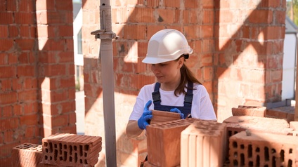 Eine Person mit weißem Helm und blauen Handschuhen steht auf einer Baustelle und hält mit beiden Händen einen roten Ziegel. Im Vordergrund liegen lose Ziegel. Im Hintergrund sieht man den Rohbau einer Mauer. 