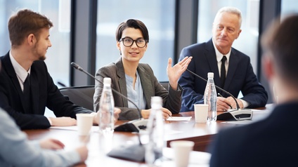 Person mit kurzen dunklen Haaren, Brille und Businesskleidung sitzt während einer Konferenz am Tisch mit mehreren anderen Personen, die aufmerksam auf die sprechende Person blicken