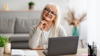 Lächelnde ältere Person mit langen blonden Haaren, Brille und Lippenstift sitzt bei einem Schreibtisch mit aufgeklapptem Laptop, Notizbuch, Stifteköcher und Zierpflanzen, im Hintergrund zeigt sich ein Wohnraum mit Sofa