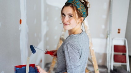 Eine Person steht mit dem Rücken zur Kamera und hat sich nach links zur Kamera aufgedreht. Die Person hält eine Malerrolle mit blauer Farbe. Im Hintergrund sind zwei Leitern