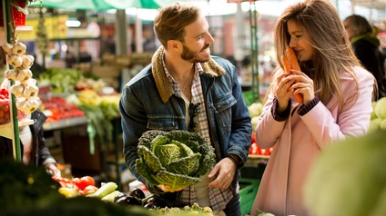 Zwei Personen stehen an Gemüsestand auf Markt, eine Person hält Kohlkopf in Händen, die andere riecht an einem Bund Karotten