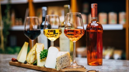 Drei Weingläser mit Rose, Weiß- und Rotwein gefüllt auf Holzbrett mit Käse und Kräutern stehend, im Hintergrund verschwommen Roseflasche