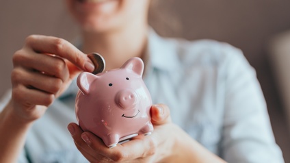 Eine Person hält ein rosa Sparschwein in der Hand und wirft eine Münze hinein.