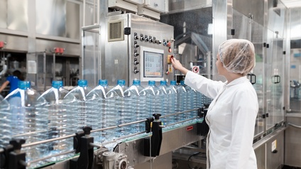 Person in weißem Kittel mit Haarnetz bedient Anlage zur Befüllung von Flaschen, vor ihr Fließband mit blauen großen Plastikflaschen