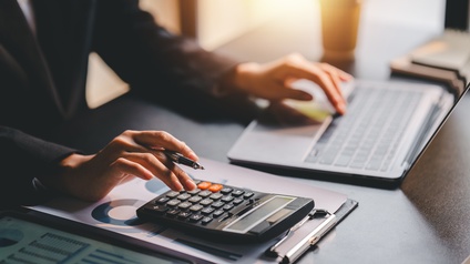 Hände einer Person in Nahaufnahme. Mit der linken Hand tippt sie auf einem Laptop, mit der rechten Hand auf einem Taschenrechner.