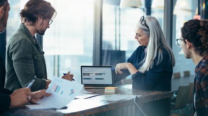 Personen an Stehtisch stehend blicken auf aufgeklappten Laptopmonitor, eine Person deutet mit Stift auf Bildschirm und spricht