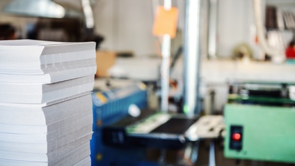 Links im Vordergrund steht ein hoher Papierstapel. Im Hintergrund steht eine Druckmaschine, bei der ein Knopf rot leuchtet