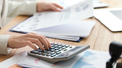 An einem Tisch sitzt eine Person in einem hellen Sakko. Die Person tippt mit der rechten Hand auf einem Taschenrechner. Darunter sind mehrere Zettel mit verschiedenen Diagrammen