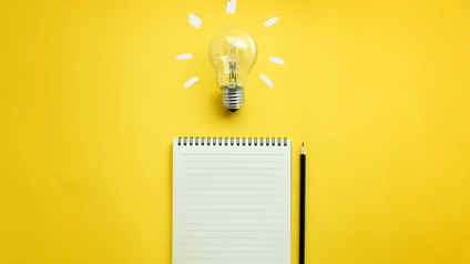 Auf gelbem Untergrund liegt ein Notizblock. Rechts neben dem Notizblock ist ein Bleistift. Über dem Notizblock ist eine Glühbirne mit weißen Strichen, die von ihr weggehen