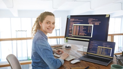 Lächelnde Personen mit Locken sitzt an Schreibtisch und hält Hände auf Tastatur, am Tisch aufgeklapptes Notebook und Computerbildschirm, auf den Monitoren Quellcodes ersichtlich