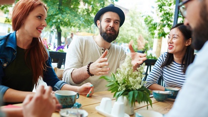 Um einen Tisch eines Gastgartens eines Lokals sitzen fünf Personen, die sich miteinander unterhalten. Auf dem Tisch sind Tassen, eine Schale mit Zucker, Salz und Pfeffer sowie eine Vase mit Blumen.