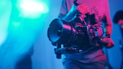 Eine Person hält mit beiden Händen eine Filmkamera vor ihren Bauch und filmt. Im Hintergrund stehen weitere Personen