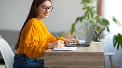 Lächelnde Person mit Billen in gelbem Pullover an Tisch vor aufgeklapptem Notebook sitzend, nimmt mit einer Hand Notizen vor und hält andere Hand auf Tastatur des Notebooks, im Hintergrund verschwommen Zimmerpflanze