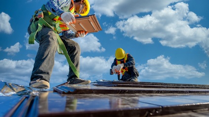 Zwei Personen mit Sicherheitswesten, Helmen und Handschuhen stehen auf einem Dach. Die Person im Vordergrund hat eine Sicherung um die Körpermitte und hält zwei Fliesen. Die Person im Hintergrund hält einen Bohrer auf das Dach