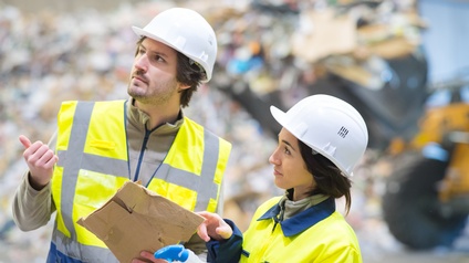 Zwei Personen in Arbeitskleidung mit Helmen stehen in einer Recyclinganlage und unterhalten sich. Sie halten eine Sprühflasche und ein Stück Altkarton in den Händen. Eine Person deutet in eine Richtung.