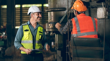 Eine Person in Rückenansicht mit Helm und Sicherheitsweste sitzt hinter einem großen Metallobjekt. In der linken Hand hält sie einen länglichen, flachen Metallstab, worauf sie blickt. Links daneben steht eine weitere Person in Schutzkleidung und lächelt.