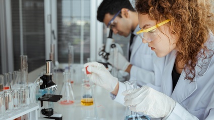 Person mit Schutzbrillen in weißem Kittel an Tisch sitzend, mit Pipette Flüssigkeit in Schälchen tropfend hält in Hand Glas mit Flüssigkeit, am Tisch Mikroskop und Röhrchen mit Flüssigkeiten, im Hintergrund weitere Person durch Mikroskop äugend
