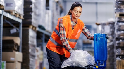 Eine Person mit geschlossenen dunklen Haaren und kariertem Hemd sowie roter Warnweste entsorgt Plastikmüll in einer blauen Tonne in einer Lagerhalle.