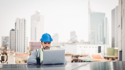 Frontale Aufnahme einer Person mit Bauhelm, die an einem Solarpanel steht und auf diesem einen aufgeklappten Laptop hat, auf dessen Bildschirm sie blickt. Im Hintergrund sind Hochhäuser