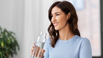 Frontale Aufnahme einer Person mit dunklen, offenen Haaren und hellblauem Pullover, die aus einem Glasbehälter Wasser trinkt. Im Hintergrund ein Fenster, eine Zimmerpflanze und weiße Vorhänge