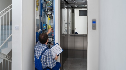 Eine Person in Arbeitskleidung hält ein Clipboard in der Hand und repariert einen Aufzug. Neben der geöffneten Lifttüre befindet sich ein elektronisches Panel, an dem die Person einen Schalter betätigt.