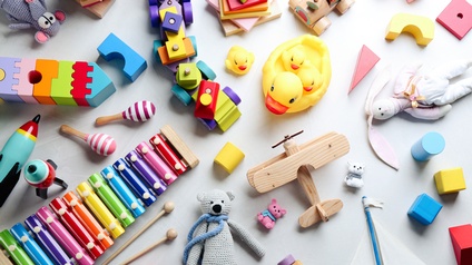 Aufnahme einer hellen Oberfläche aus der Vogelperspektive, auf der verschiedene Spielsachen wie Bauklötze, ein Holzzug, Rasseln, Stofftiere, eine Xylophon, Gummienten, ein Segelboot und ein Holzflugzeug sind