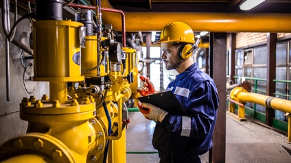 Seitliche Aufnahme einer Person mit gelbem Helm, Kopfhörern, Overall, Handschuhen und Klemmbrett, die neben dicken, vor einer Wand stehenden gelben Rohren steht und darauf blickt